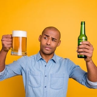 Ein Mann schaut skeptisch auf ein Glas Bier und eine Flasche Bier in seiner Hand. Ein alkoholfreies Getränk kann auch Alkohol enthalten.