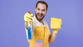 Ein Mann in Hausarbeitskleidung hält eine Sprühflasche ins Bild | Symbolbild: Höhenflug im Haushalt: Männer überschätzen ihre Hausarbeit