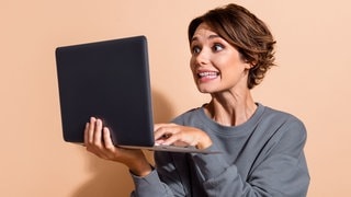 Frau schaut überrascht auf ihr Laptop. | Symbolbild für häufige Schreibfehler in E-Mails und die richtige Lösung laut Duden für Grußformeln und Kommasetzung.