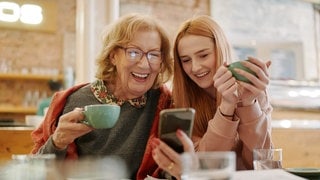 Zwei Frauen aus unterschiedlichen Generationen schauen lachend auf ein Handy in einem Café. Symbolbild für typische Wörter, die vor allem Boomer benutzen – und in der Sprache anderen Alters eher keine Rolle spielen.