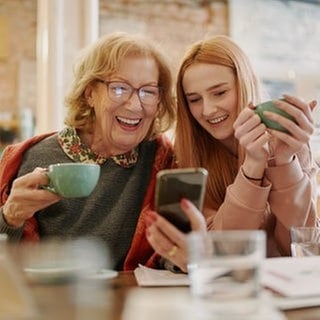 Zwei Frauen aus unterschiedlichen Generationen schauen lachend auf ein Handy in einem Café. Symbolbild für typische Wörter, die vor allem Boomer benutzen – und in der Sprache anderen Alters eher keine Rolle spielen.