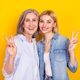 Boomer-Mutter und jüngere Tochter lächelnd nebeneinander mit „Peace“-Handzeichen. Symbolbild für typische Wörter, die vor allem Boomer benutzen – und in der Sprache anderer Generationen eher keine Rolle spielen.