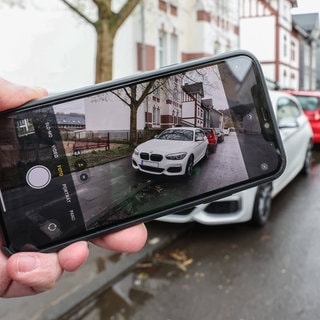 Eine Hand mit einem Smartphone fotografiert einen vermutlichen Falschparker um diesen anzuzeigen. | Symbolbild: Falschparker melden