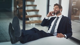 Symbolbild: Junger Mann im Anzug bei einem privaten Gespräch an seinem Handy am Arbeitsplatz.