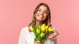 Junge Frau mit einem Strauß Blumen in der Hand lacht. Symbolbild für Sprüche, mit denen wir es durch die Blume sagen können, offiziell freundlich.