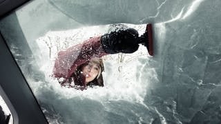 Junge Frau mit Enteiser, sie kratzt die verschneite und gefrorene Scheibe des Autos im Winter frei.