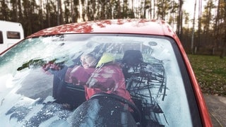 Die Autoscheibe ist im Winter auch von innen gefroren und vereist. Eine Frau, dick eingepackt in Winterkleidung, kratzt die Scheibe von innen frei.