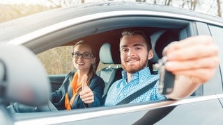 Ein junger Mann am Steuer eines Autos während seiner Führerscheinprüfung. Die Prüferin signalisiert ihm, dass er den Test bestanden hat.