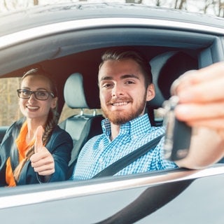 Ein junger Mann am Steuer eines Autos während seiner Führerscheinprüfung. Die Prüferin signalisiert ihm, dass er den Test bestanden hat.