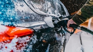 Ein schwarzes Auto wird im Winter an einer Tankstelle betankt. Es fallen Schneeflocken. Das Foto soll den erhöhten Spritverbrauch im Winter, beispielsweise auf dem Weg in den Skiurlaub, verdeutlichen. Wir klären, woran das liegt und welche Tipps es gibt, um die größtens Spritfresser im Winter zu vermeiden.