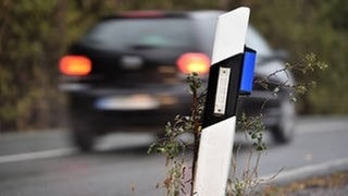 Auto fährt über eine Straße. Am Straßenrand steht ein Leitpfosten mit blauen Reflektoren, ein Wildwarnreflektor. Symbolbild für Artikel zum Verkehrszeichen und dessen Bedeutung.