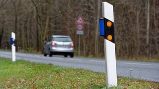 Auto fährt über eine Straße. Am Straßenrand steht ein Leitpfosten mit blauen Reflektoren, ein Wildwarnreflektor. Symbolbild für Artikel zum Verkehrszeichen und dessen Bedeutung für Autofahrer.