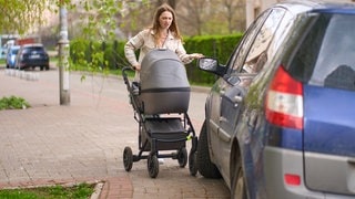 Frau spaziert mit Kinderwagen auf Gehweg und wird von geparkten Auto blockiert | Symbolbild für parken auf Gehweg: Wann gibt es Bußgelb und Punkt in Flensburg