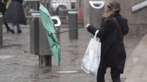 Frau bei Sturm mit einem Regenschirm - am Wochenende soll es wegen Bomben-Zyklon in BW und RLP wärmer und stürmisch werden