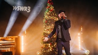 Mark Forster mit Cap und Mikro auf der Bühne vor einem beleuchteten Weihnachtsbaum