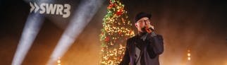 Mark Forster mit Cap und Mikro auf der Bühne vor einem beleuchteten Weihnachtsbaum