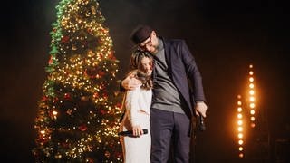 Annisa und Mark Forster auf der Bühne vor einem Weihnachtsbaum, die beiden umarmen sich und sehen glücklich aus