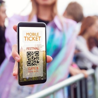 Junge Frau hält ein Smartphone in der Hand mit einem mobilen Ticket, im Hintergrund Konzert-Stimmung.