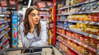 Eine Frau steht mit ihrem Wagen im Supermarkt und schaut frustriert die Regale an