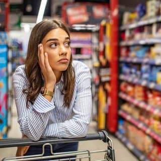 Eine Frau steht mit ihrem Wagen im Supermarkt und schaut frustriert die Regale an