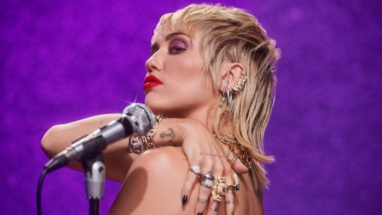Die Sängerin Miley Cyrus vor einer lila Wand mit einem silbernen Mikrofon. 
