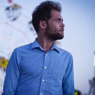 Der Singer-Songwriter Passenger trägt ein weiß-blau gestreiftes Hemd und steht vor einem Riesenrad, sein größter Hit ist „Let Her Go“