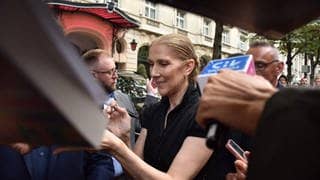 Celine Dion stoi przed hotelem w Paryżu w czarnej sukni i uczesanych włosach i rozdaje autografy