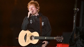 Ed Sheeran spricht auf der Bühne in ein Mikrofon, während er eine Akustikgitarre umhängen hat