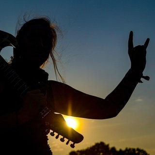 Frau mit E-Gitarre auf der Schulter zeigt den Metalgruß, die Pommesgabel.