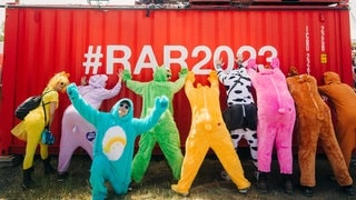 Eine Gruppe steht in einer Reihe verkleidet mit bunten Kostümen an einem Container bei Rock am Ring mit dem Rücken zur Kamera