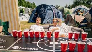 Zwei Männer sitzen hinter einem Bier-Pong-Tisch auf einem Sofa bei Rock am Ring auf dem Campingplatz