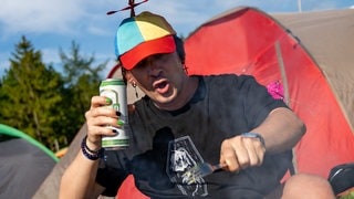 Ein junger Mann sitzt auf einem Campingstuhl mit einem kleinen Sonnenschirm-Hut auf dem Kopf vor Zelten bei Rock am Ring