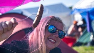 Eine junge Frau hält eine gegrillte Wurst hoch und sitzt auf einem Campingstuhl bei Rock am Ring