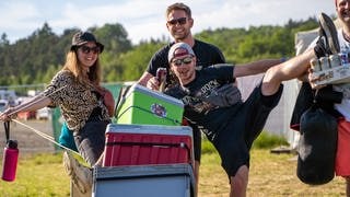 Eine junge Frau und drei Männer sind beladen mit Bierdosen und Kühlboxen auf dem Campingplatz von Rock am Ring unterwegs