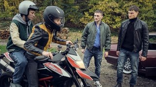 Tatort-Kritik Stuttgart „Zerrissen“: David und Sozialarbeiterin Aro auf einem Motorrad. Neben an stehen Davids Cousins und schauen aggressiv.