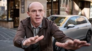 Hans Drescher (Gerhard Liebmann) steht im Tatort aus Franken (6.10.) auf einer Straße, beide Arme hat er ausgestreckt.