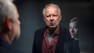 Szenenbilder aus dem Tatort „Borowski und das ewige Meer“. Borowski blickt durch eine verspiegelte Scheibe, erkennt sich darin selbst und das Gesicht einer jungen Frau. Tatort-Checker Michael Haas spricht in seiner Kritik von einem „gechillten und zarten“ Krimi, obwohl es gleich eine ganze Mordserie gibt!