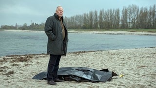 Szenenbilder aus dem Tatort „Borowski und das ewige Meer“. Der Kommissar steht am Meerestrand neben einem schwarzen Leichensack Tatort-Checker Michael Haas spricht in seiner Kritik von einem „gechillten und zarten“ Krimi, obwohl es gleich eine ganze Mordserie gibt!