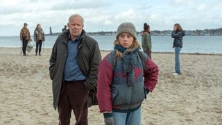 Szenenbilder aus dem Tatort „Borowski und das ewige Meer“. Kommissar Borowski steht mit einer jungen Klimaaktivistin am Strand. Tatort-Checker Michael Haas spricht in seiner Kritik von einem „gechillten und zarten“ Krimi, obwohl es gleich eine ganze Mordserie gibt!