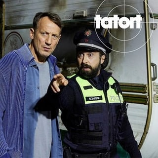 Auf einem Klostergelände ist ein Wohnwagen abgebrannt – es gab einen Toten. Kommissar Falke und ein Polizist sind am Tatort. Szenenbild aus dem Hamburger Tatort „Schweigen“ mit Kommissar Falke.