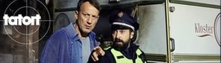 Auf einem Klostergelände ist ein Wohnwagen abgebrannt – es gab einen Toten. Kommissar Falke und ein Polizist sind am Tatort. Szenenbild aus dem Hamburger Tatort „Schweigen“ mit Kommissar Falke.